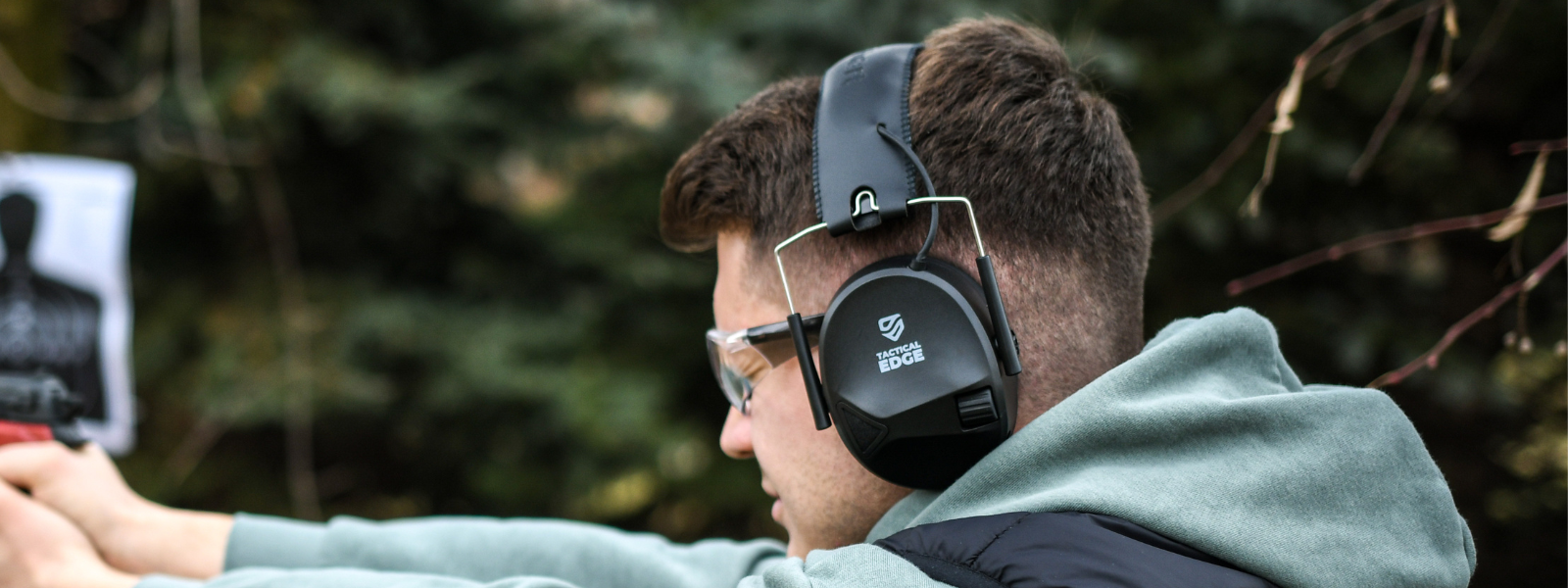 Shooter wearing TradeSmart Safety TacticalEdge electronic earmuffs and safety glasses while aiming at a target outdoors.