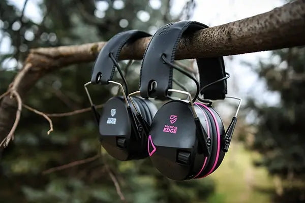 Pair of TradeSmart Safety TacticalEdge electronic earmuffs, black and black with pink accents, hanging on a tree branch outdoors